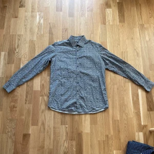 Mönstrad skjorta från Sand - Säljer en snygg mönstrad skjorta från Sand i storlek 43. Skjortan har ett unikt blommigt mönster i grått och vitt, perfekt för både vardag och fest. Den är långärmad och har knappar hela vägen ner. Materialet känns mjukt och bekvämt, perfekt för alla säsonger.