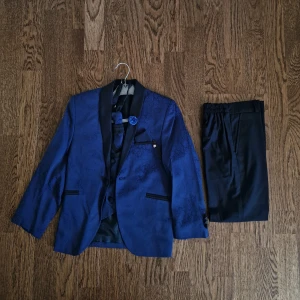 Blå kostym med matchande väst, slips och byxor för barn - Elegant blå kostym för pojkar, perfekt för festliga tillfällen! Setet innehåller kavaj, mönstrad väst, slips och svarta byxor. Fint skick, använd endast några gånger.  Pris: 599 kr. 