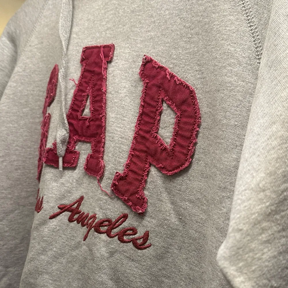 Säljer en grå hoodie från GAP med röd text 'Los Angeles' på framsidan. Den är supermjuk och perfekt för kyliga dagar. Har en stor ficka fram och en justerbar huva. Perfekt för en avslappnad stil eller mysiga hemmakvällar. Står M på tröjan mena skulle snarare säga xs. Tröjor & Koftor.