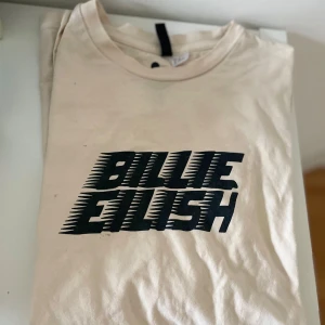 Beige Billie Eilish t-shirt från H&M - Säljer en beige t-shirt från H&M med Billie Eilish-tryck i svart. Den är i bomull och har en normal passform. Perfekt för fans av Billie Eilish! 🖤