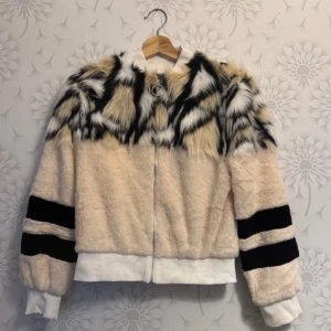 Beige och svart pälsjacka - Säljer en unik beige pälsjacka med svart och vit mönstrad detalj på axlarna. Jackan har långa ärmar med svarta ränder och en dragkedja framtill. Perfekt för att sticka ut i mängden!