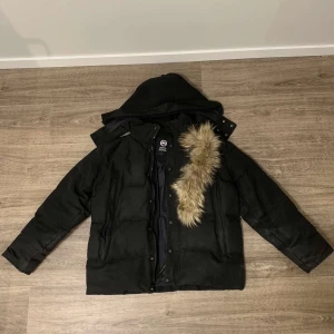 Svart dunjacka från Canada Goose - Säljer en svart dunjacka från Canada Goose med avtagbar päls på huvan. Jackan har en broderad logga på ärmen och stängs med dragkedja och knappar. Perfekt för kalla vinterdagar.