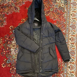 Svart dunjacka från Everest - Säljer en svart dunjacka från Everest med stor huva och dragkedja. Jackan har en långärmad design och är perfekt för kalla dagar. Den har praktiska fickor och en stilren look. Köptes från Stadium för några år sedan. Som ses på bilden är ena dragskon borta, men det går att fixa!