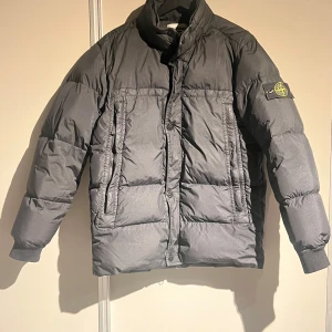 Svart dunjacka från Stone Island - Säljer en grå dunjacka från Stone Island med en unik crinkle-effekt. Jackan har en hög krage och stängs med både dragkedja och knappar. Den är tillverkad i ett lätt nylonmaterial och har märkets ikoniska logga på ärmen.