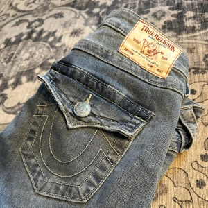 Blå jeans från True Religion storlek 26  - Snygga blå jeans från True Religion med klassiska bakfickor och logotypdetaljer. Jeansen har en bootcut-stil och är perfekta för en avslappnad look. De har en knapp och dragkedja framtill. Låg modell samt upplagda. Passar mig i längd som är 157cm. 