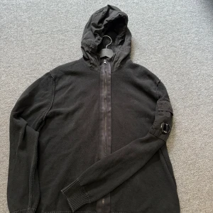 Svart jacka med huva från C.P. Company - Säljer en svart jacka med huva från C.P. Company. Jackan har en dragkedja framtill och en unik detalj på ärmen. Perfekt för en stilren look.