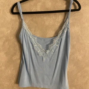 Ljusblå linnetopp från DeeZee - Säljer en söt ljusblå linnetopp från DeeZee med spetsdetaljer vid halsringningen. Toppen har smala axelband och är perfekt för varma dagar. Materialet är mjukt och stretchigt för en bekväm passform. 🌸
