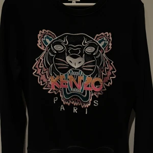 Svart tröja med tigerbroderi från Kenzo - Säljer en svart tröja från Kenzo med ett stort, färgglatt tigerbroderi på framsidan. Tröjan har långa ärmar och en rund halsringning. Perfekt för att ge din outfit en cool touch! Köpt här på plick men kommer ej till användning längre🩷