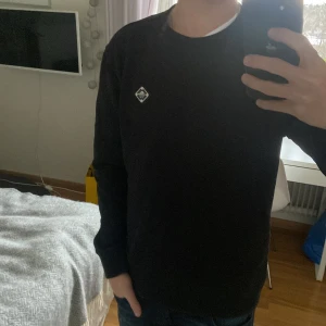 J.lindberg tröja - Säljer en stilren svart tröja med rund hals och en liten logotyp på bröstet. Tröjan har långa ärmar och är perfekt för en avslappnad look.