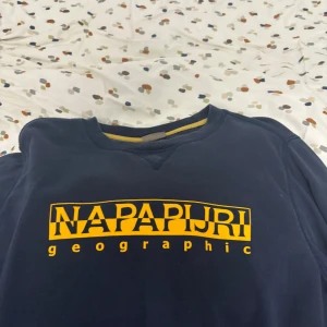 Mörkblå sweatshirt från Napapijri - Säljer en snygg mörkblå sweatshirt från Napapijri med gult tryck på bröstet och ett coolt märke på ärmen. Perfekt för en avslappnad stil.