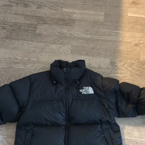 Svart dunjacka från The North Face - Säljer en svart dunjacka från The North Face. Jackan har en pufferdesign med dragkedja och hög krage för extra värme. Perfekt för kalla dagar.