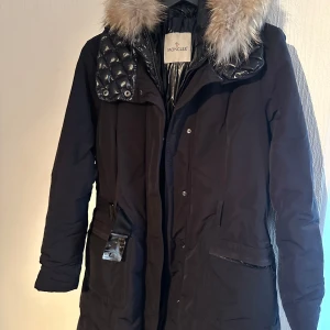 Svart vinterjacka från Moncler - Snygg svart vinterjacka från Moncler med quiltade detaljer och en avtagbar huva med päls. Jackan har ett bälte i midjan och stängs med dragkedja och knappar. Perfekt för kalla dagar.