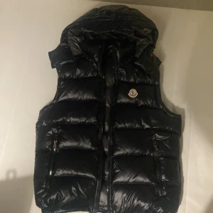 Svart dunväst från Moncler - Snygg svart dunväst från Moncler med dragkedja och huva. Perfekt för kyligare dagar och ger en stilren look. Västen har en glansig finish och två praktiska fickor framtill. Nfc funkar och kvittot finns också. Och den passar som M. Priset kan diskuteras.
