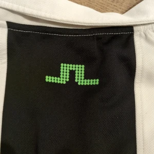 Vit och svart pikétröja från J.Lindeberg - Snygg vit och svart pikétröja från J.Lindeberg med korta ärmar och dragkedja framtill. Tröjan har en stilren design med ett grönt märke på ryggen. Perfekt för en sportig look!