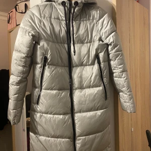 Grå lång dunjacka från Carry VNG - Säljer en lång och varm grå dunjacka från Carry VNG Ladies Collection. Jackan har en huva och dragkedja framtill samt två praktiska fickor med dragkedjor. Perfekt för kalla dagar. Köpte den i polen för 4000kr men har knappt kommit till användning❤️