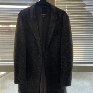 Mörkgrå kavaj från SIR of Sweden - Elegant mörkgrå kavaj från SIR of Sweden med diskret mönster. Kavajen har en klassisk design med knappar framtill och innerfickor. Perfekt för en stilren look.