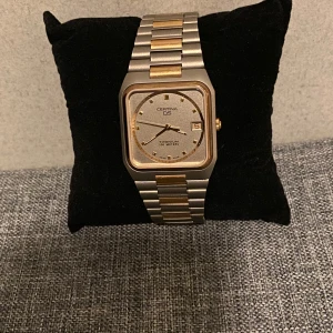 Certina DS  i silver och guld - Elegant Certina DS herrklocka med en stilren design. Klockan har en fyrkantig urtavla i silver med guldiga detaljer och datumvisning. Armbandet är i rostfritt stål med guldiga inslag, vilket ger en lyxig känsla.