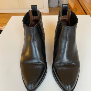Svarta Rodebjer läderboots med klack - Snygga svarta Rodebjer boots i skinn med spetsig tå och klack i mycket bra skick. De har en stilren design med elastiska paneler på sidorna för enkel på- och avtagning. Gummisula. Baksidan har ett diskret ormskinnsmönster som ger en extra touch.
