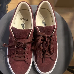 Vinröda sneakers från Polo Ralph Lauren - Snygga vinröda sneakers från Polo Ralph Lauren i mocka med klassisk snörning. Skorna har en vit sula och det ikoniska Polo-logotypen broderad på sidan. Perfekta för en stilren look.