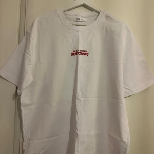 Vit t-shirt från Samsøe Samsøe - Vit t-shirt från Samsøe Samsøe med tryck fram och bak. Framsidan har texten 'Samsøe Samsøe Gone Fishing' i rött. Baksidan pryds av en stor röd ros och texten 'Please Stand By Samsøe Samsøe Gone Fishing'. Perfekt för en avslappnad stil. Lite oversized