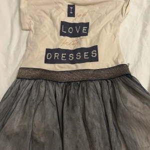 Beige och mörkgrå klänning från Little Pomp de Lux - Charmig klänning från Little Pomp de Lux med texten 'I just love pretty dresses' på framsidan. Överdelen är veige med korta ärmar och kjolen är mörkgrå och luftig med ett glittrigt midjeband. Perfekt för små fashionistas! Den kommer tvättas och strykas innan den skickas! ⭐️