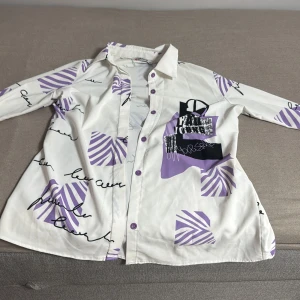 Vit och lila skjorta med grafiskt mönster - Snygg vit skjorta med lila och svarta grafiska mönster och textdetaljer. Skjortan har långa ärmar och knappar framtill. Perfekt för en unik och trendig stil.