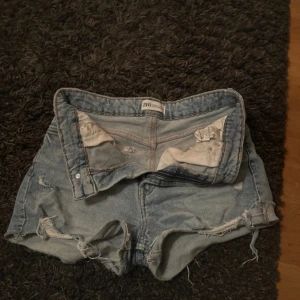 Blå jeansshorts från Zara - Säljer ett par blå jeansshorts från Zara med slitna detaljer och råa kanter. Shortsen har en klassisk femficksdesign och är perfekta för en avslappnad stil. De är tillverkade i ett robust denimtyg.
