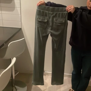 Grå velourbyxor från Juicy Couture - Säljer ett par gröna  velourbyxor från Juicy Couture i storlek XS. Byxorna har en bekväm passform med resår i midjan och snörning framtill. De har två bakfickor med knappar som ger en snygg detalj.Saknas dock knapparna på banden se sista bilden säljer då jag har vuxit ur dom