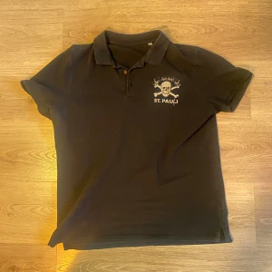 St. Pauli piké - Snygg svart pikétröja med krage och knappar. Har ett coolt St. Pauli-tryck med dödskalle och texten 'Sylt Boyz' på bröstet. Perfekt för en avslappnad stil.