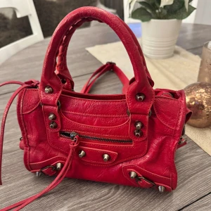 Röd handväska med nitar - Snygg röd handväska i skinn med coola nitar och dragkedja framtill. Väskan har flätade handtag och en avtagbar axelrem för extra stil och funktionalitet. Perfekt för att ge din outfit en färgklick!