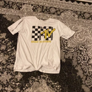 Vit MTV t-shirt - Säljer en cool vit t-shirt med MTV-logga i svart och gult. T-shirten har ett schackrutigt mönster bakom loggan och är kortärmad. Perfekt för alla som älskar retrostil och musik.