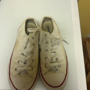 Vita sneakers från Converse - Säljer ett par klassiska vita Converse sneakers med snörning och röd rand längs sulan. Perfekta för en avslappnad stil. Obs, behöver rengöras. 