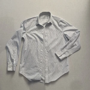 Vit och blå randig skjorta från Calvin Klein - Snygg vit skjorta från Calvin Klein med smala blå ränder och små prickar. Skjortan har en slim fit passform och är tillverkad i ett non-iron material för enkel skötsel. Perfekt för en stilren look.
