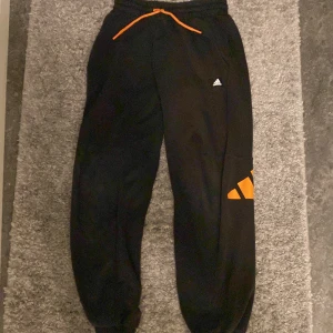 Svarta mjukisbyxor från Adidas - Säljer ett par svarta mjukisbyxor från Adidas med orangea detaljer och snörning i midjan. Byxorna har en avslappnad passform och är perfekta för en bekväm stil.