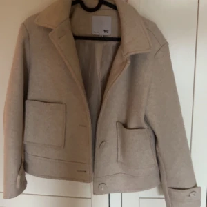Beige kappa från 157 - Säljer en stilren beige kappa från 157 med stora fickor och knappar framtill. Jackan har en klassisk krage och är perfekt för kyligare dagar. Den har en rak passform och är tillverkad i ett mjukt material.