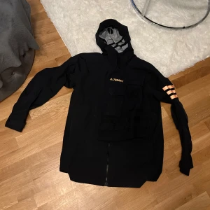 Svart jacka från Terrex - Säljer en svart jacka från Terrex med huva och dragkedja. Jackan har långa ärmar och en praktisk ficka på bröstet. Perfekt för utomhusaktiviteter och har en sportig look med orange detaljer på ärmen.