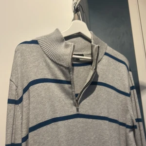 Grå randig tröja från Dressmann - Säljer en grå tröja från Dressmann med blå ränder och dragkedja vid halsen. Tröjan har en ribbad krage och är perfekt för en avslappnad stil.