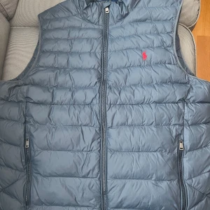 Blå dunväst från Polo Ralph Lauren - Säljer en snygg blå dunväst från Polo Ralph Lauren med dragkedja och broderad logga i rött på bröstet. Perfekt för kyligare dagar och ger en stilren look. Västen har två praktiska fickor med dragkedja.