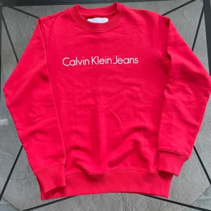 Röd sweatshirt från Calvin Klein Jeans - Säljer en snygg röd sweatshirt från Calvin Klein Jeans med deras ikoniska logga tryckt på bröstet. Tröjan har långa ärmar och ribbade muddar vid ärmslut och nederkant. Perfekt för en casual look!