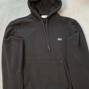 Svart hoodie från Lacoste - Säljer en klassisk svart hoodie från Lacoste med den ikoniska krokodilloggan på bröstet. Tröjan har en justerbar huva med dragsko och en praktisk magficka. Perfekt för en stilren och bekväm look. Vid frågor är det bara att höra av er.