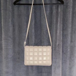 Beige axelväska - Beige axelväska med design. Väskan har en lång rem som gör den enkel att bära över axeln. Knapp som öppning till väskan