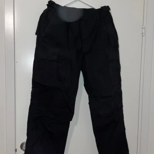 Svarta cargopants - Säljer ett par svarta cargopants med flera fickor och justerbara remmar i midjan. Perfekta för en avslappnad och funktionell stil. Byxorna har en loose passform och är tillverkade i ett slitstarkt material. *EJ DENIM*