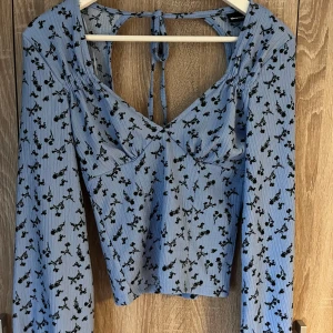 Blommig blå topp från Gina Tricot - Säljer en söt blå topp från Gina Tricot med svart blommönster. Toppen har långa ärmar och en öppen rygg med knytband. 