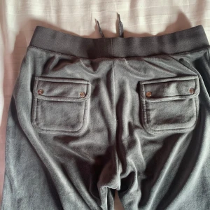 Grå mjukisbyxor från Juicy Couture - Snygga grå mjukisbyxor från Juicy Couture med snörning i midjan och praktiska fickor med knappar bak. Perfekta för en avslappnad stil.