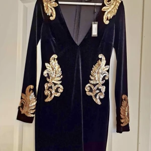 Svart sammet klänning med guld broderi - Elegant svart sammet klänning från Goddiva med v-ringning och långa ärmar. Klänningen har vackra guld broderade detaljer på axlar, ärmar och midja. Perfekt för en glamorös kväll.