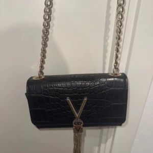 Svart valentino axelväska med kedja - Snygg svart axelväska i skinn med krokodilpräglat mönster och en elegant kedja som axelrem. Väskan har en dekorativ tofsdetalj framtill och en stilren design som passar perfekt för en chic look.