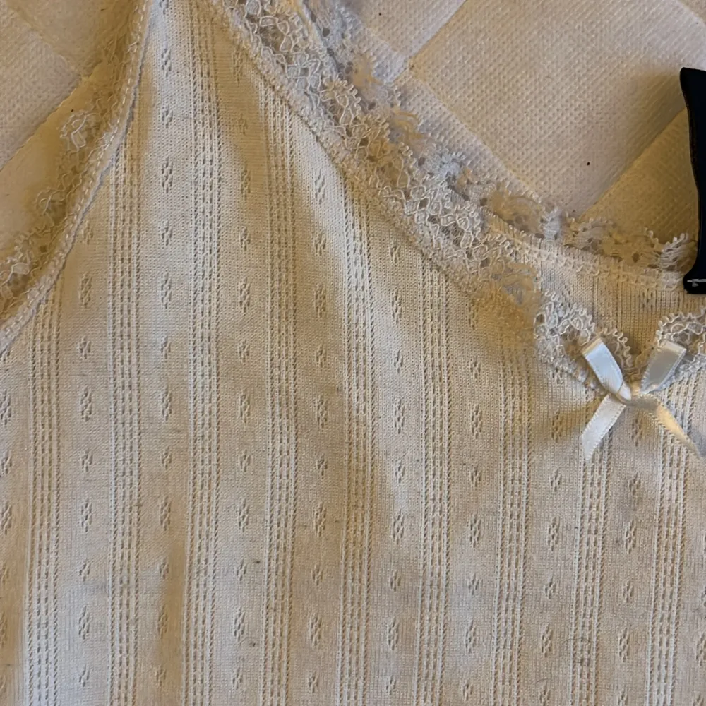 Säljer en söt vit linnetopp från H&M Divided med spetsdetaljer och en liten rosett framtill. Toppen har smala axelband och ett diskret mönster i tyget. Perfekt för varma dagar eller som ett lager under en tröja.. Yläosat.