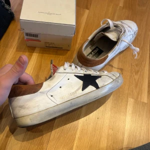 Golden goose - Snygga vita sneakers från Golden Goose med en ikonisk svart stjärna på sidan. Skorna har bruna detaljer vid hälen och vita snören. Perfekta för en avslappnad stil med en touch av lyx.