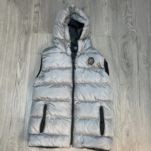 Grå dunväst från Philipp Plein - Snygg grå dunväst från Philipp Plein med huva och dragkedja. Västen har en cool logga på bröstet och svarta detaljer vid fickorna. Perfekt för att hålla stilen när det är kyligt ute.