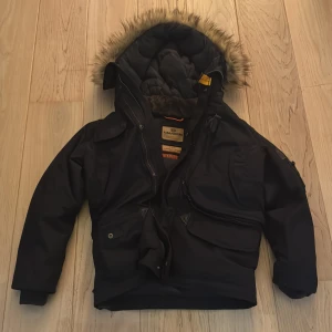 Svart vinterjacka från Parajumpers - Säljer en svart vinterjacka från Parajumpers med pälsfodrad huva och flera praktiska fickor. Jackan har en snygg patch på ärmen och dragkedja framtill. Perfekt för kalla dagar!